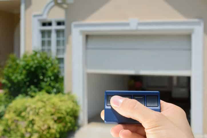 A hand holding a remote control opens a white garage door attached to a beige house with plants nearby. | M&M Garage Doors