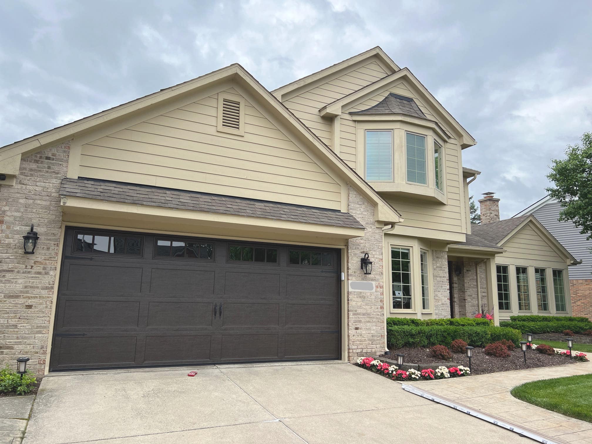M&M_garage_doors_replacement Two-story suburban house with tan siding and brick exterior, dark garage door, landscaped front yard, and overcast sky.