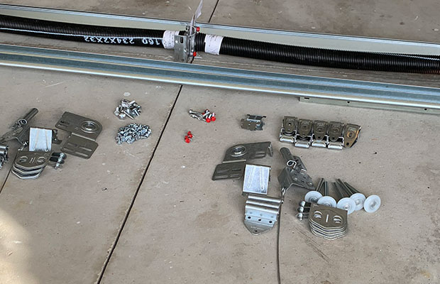 garage-door-parts-forest-hills-mi Assorted metal hardware, bolts, brackets, and rollers are laid out on a concrete floor near a garage door track and torsion spring.
