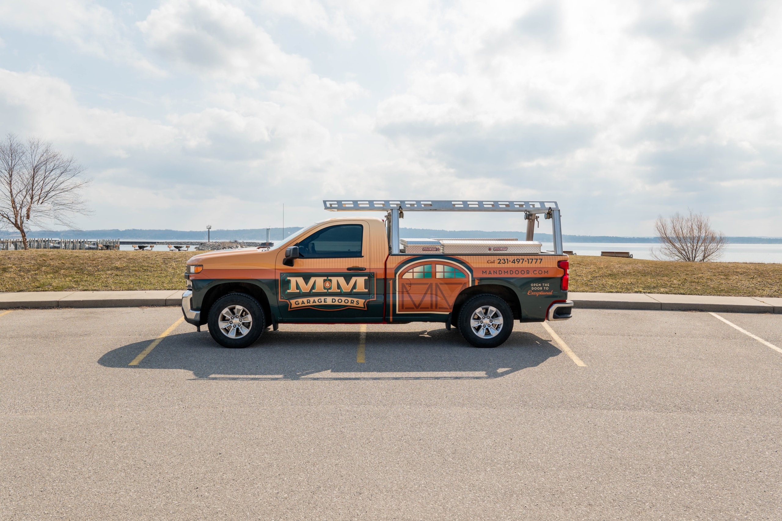 TC13 A parked orange and tan pickup truck with a ladder rack, displaying "M&M Garage Doors" branding and a phone number on the side, in a lot near a body of water.