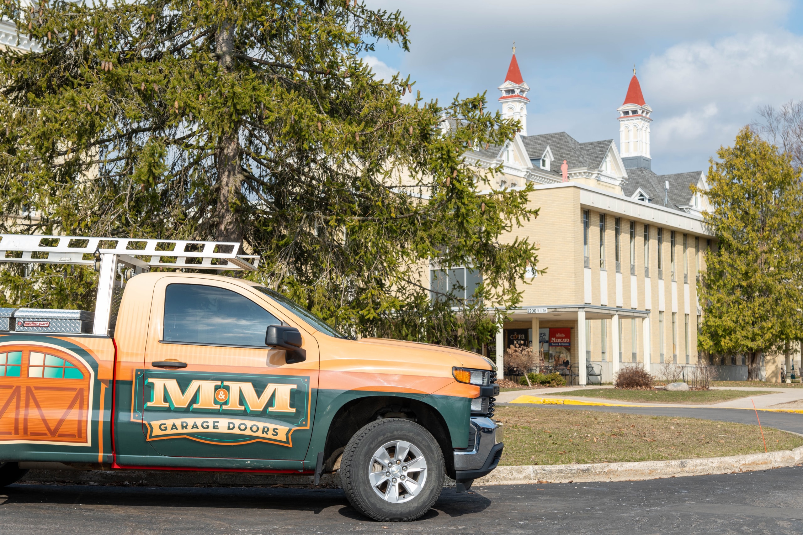 TC7 A pickup truck with an "M&M Garage Doors" logo is parked in front of a light-colored building with red-roofed towers and a tree nearby.