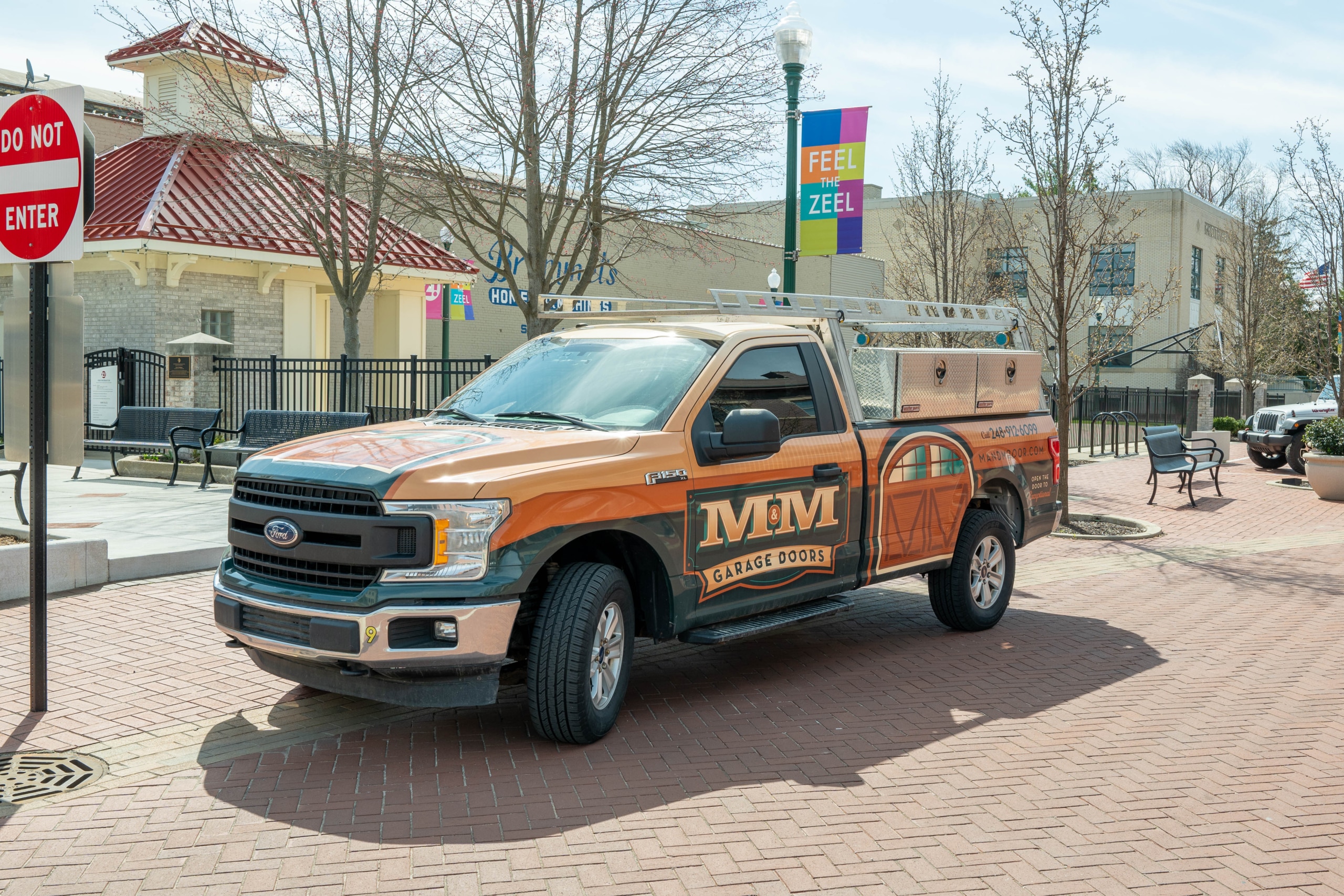 A brown Ford F-150 truck with "MM Garage Doors" branding is parked on a brick street near a no entry sign and some buildings.