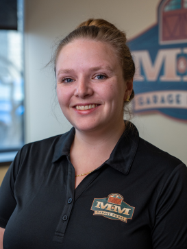 A woman in a black M&M Garage Doors polo shirt smiles at the camera, standing indoors with a company logo visible on the wall behind her. | M&M Garage Doors