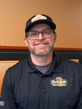 Man with glasses, short beard, and hat, wearing a black polo shirt with "MM" logo, standing indoors against a tan and orange background. | M&M Garage Doors