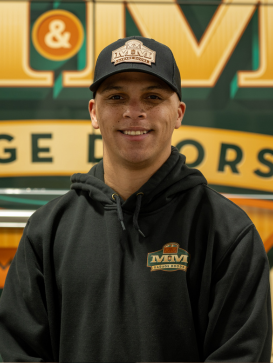 A person wearing a black hoodie and black cap with an MVM Garage Doors logo stands in front of an MVM Garage Doors sign. | M&M Garage Doors