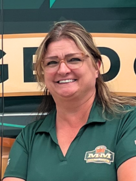 A woman wearing glasses and a green MM polo shirt stands in front of a sign with large yellow and green letters. | M&M Garage Doors