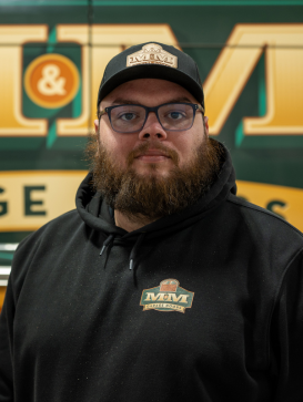 A bearded person wearing glasses, a black hoodie, and a black cap with "M&M Autobody" logos, stands in front of a green and yellow "M&M Autobody" sign. | M&M Garage Doors