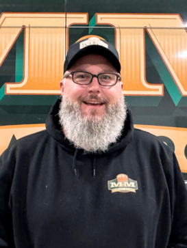 A bearded man wearing glasses, a black hoodie, and a cap stands in front of a large green and yellow sign with "MM" visible. | M&M Garage Doors