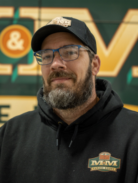 Man with a beard and glasses wears a black hoodie and cap, both with "M&M Garage Doors" logos, standing in front of a sign with large yellow letters. | M&M Garage Doors