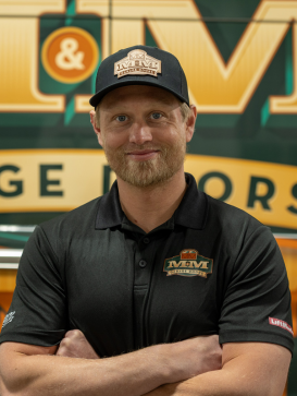 Man in black M&M Garage Motors cap and polo shirt stands with arms crossed in front of a branded sign. | M&M Garage Doors
