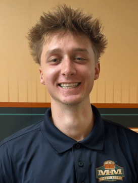 Young adult with spiky hair smiles at the camera, wearing a dark polo shirt with a logo that reads "M&M Garage Doors" against a beige and green background. | M&M Garage Doors