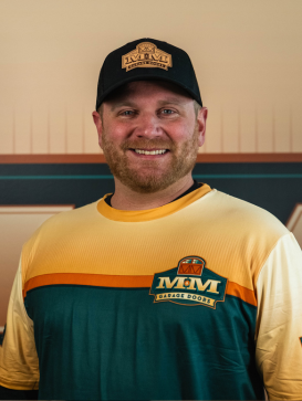 A man wearing a black cap and a yellow and green IVW Garage Doors shirt stands smiling in front of a matching background. | M&M Garage Doors