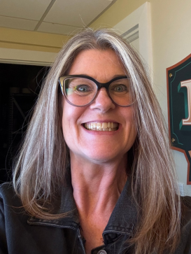 A woman with long gray hair and glasses smiles widely at the camera, wearing a dark jacket indoors. | M&M Garage Doors