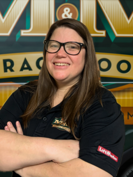 A woman with long brown hair and glasses, wearing a black polo shirt with embroidered logos, stands smiling with her arms crossed in front of a colorful sign. | M&M Garage Doors