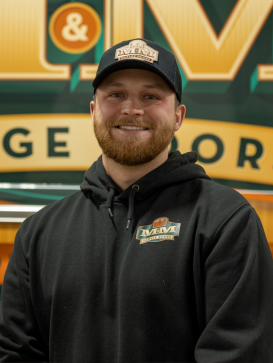 A man wearing a black hoodie and cap with matching logos stands indoors, smiling in front of a sign with large yellow and green lettering. | M&M Garage Doors