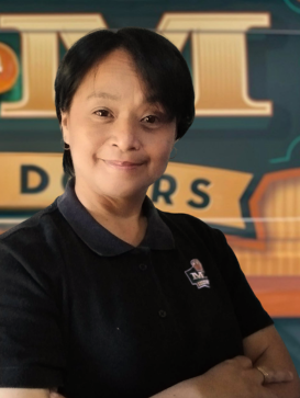 Person with short black hair and a black collared shirt stands in front of a sign with large letters and an orange graphic, smiling with arms crossed. | M&M Garage Doors