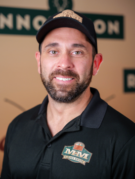 Man with a beard wearing a black baseball cap and a black polo shirt with an "MM Draft Door" logo, standing in front of a blurred sign background. | M&M Garage Doors