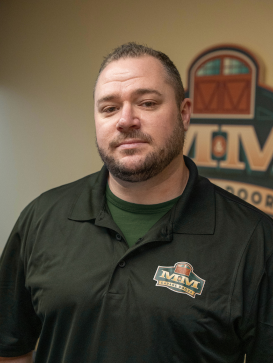 Man with short hair and beard wearing a black M&M Exteriors polo shirt stands indoors in front of an M&M Exteriors logo sign. | M&M Garage Doors