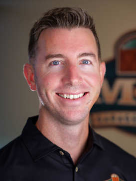 A man with short brown hair and blue eyes smiles at the camera, wearing a black collared shirt. A blurred logo is visible in the background. | M&M Garage Doors