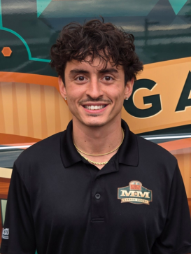 A young man with curly hair and a mustache smiles at the camera, wearing a black polo shirt with an "M&M" logo, standing in front of a colorful background. | M&M Garage Doors