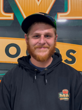 A man with a red beard and black hoodie stands in front of a large sign with yellow and green letters. | M&M Garage Doors