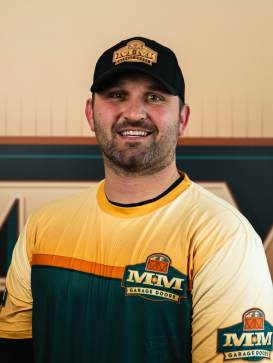 A man in an Mm Garage Doors shirt and hat stands smiling in front of a matching Mm Garage Doors logo backdrop. | M&M Garage Doors