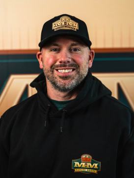 A man in a black hoodie and cap with matching logos smiles at the camera, standing in front of a similarly branded background. | M&M Garage Doors
