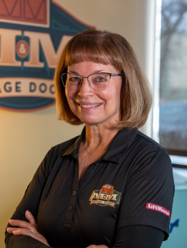 Woman with short brown hair and glasses, wearing a black collared shirt with embroidered company logos, stands smiling with arms crossed in an office setting. | M&M Garage Doors